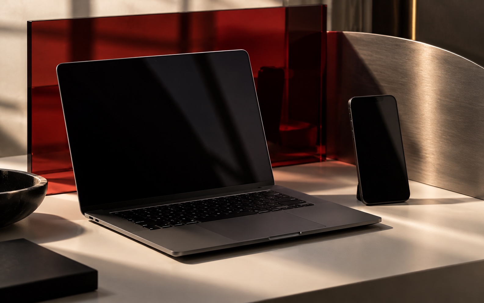 暖白桌面上的黑色笔记本电脑和手机，背景为深红玻璃与金属曲面